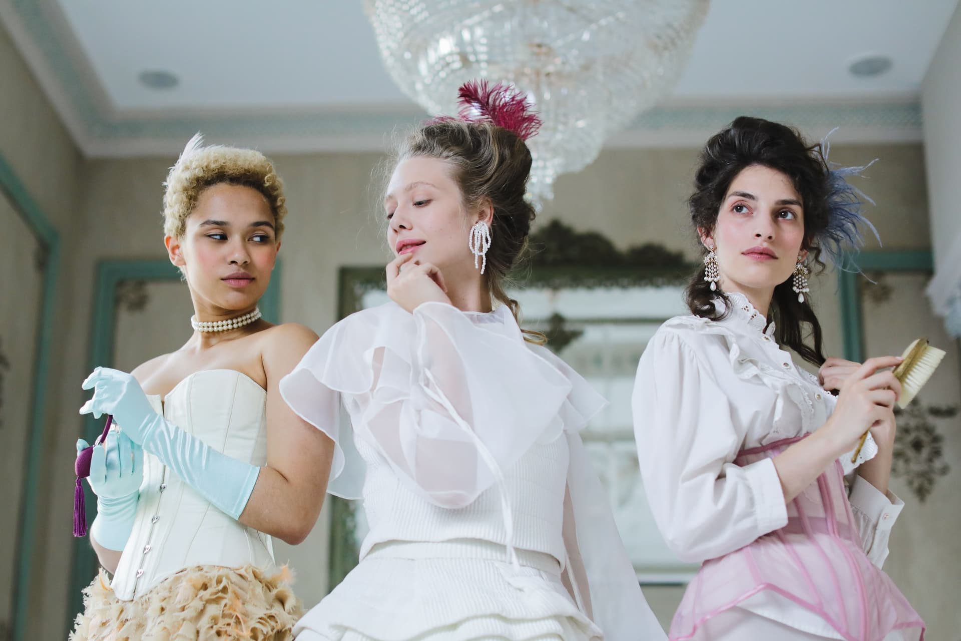 Three models in Rococo-inspired couture prepare in an ornate salon with a crystal chandelier