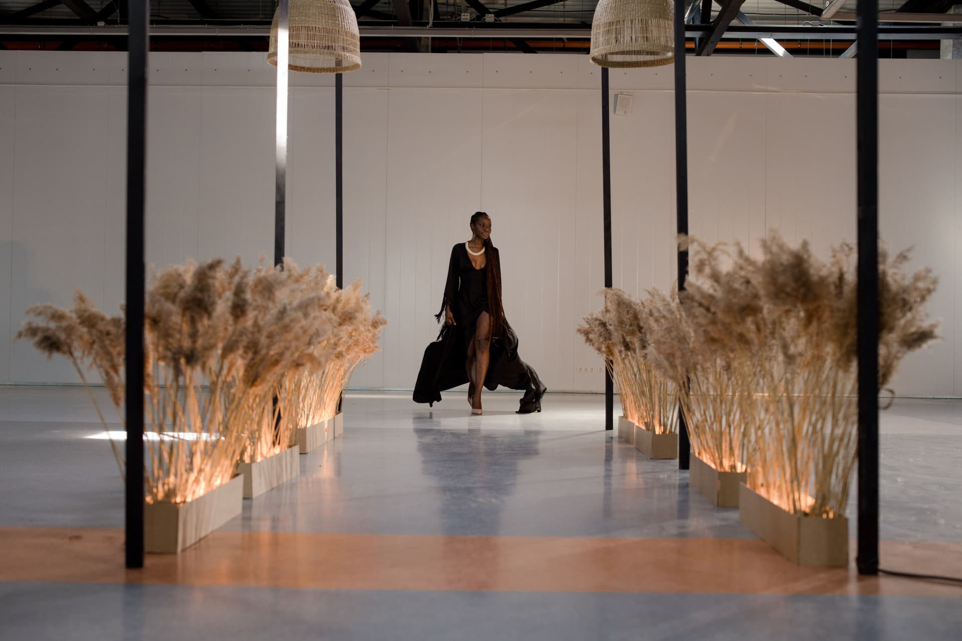 A model in a long black dress walks a runway lined with illuminated dried plants