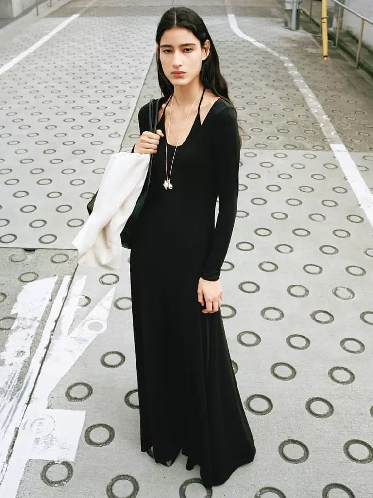 A model in a long black dress carrying a cream coat against an urban street
