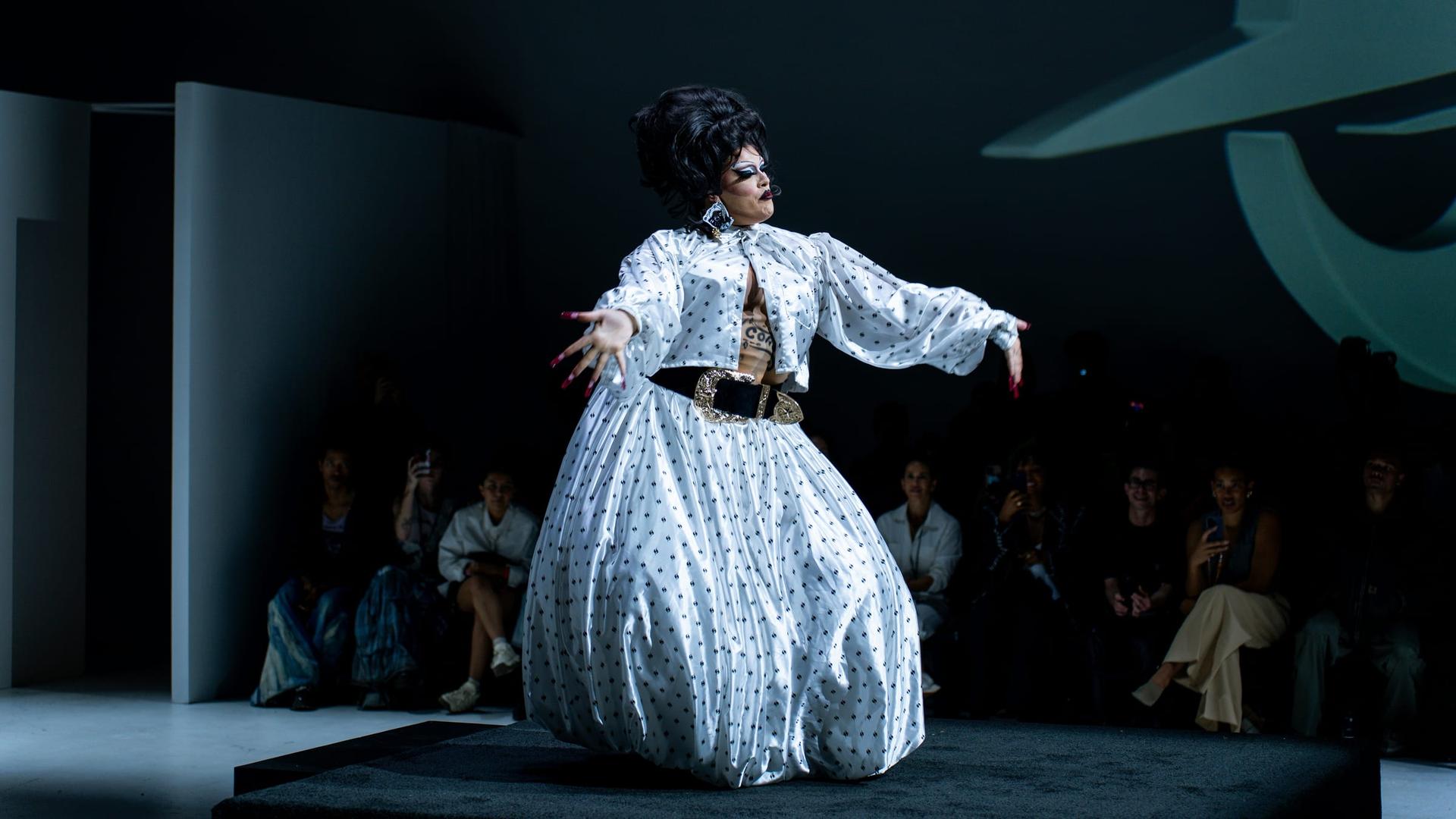 A performer in patterned couture strikes a pose on a dimly lit runway
