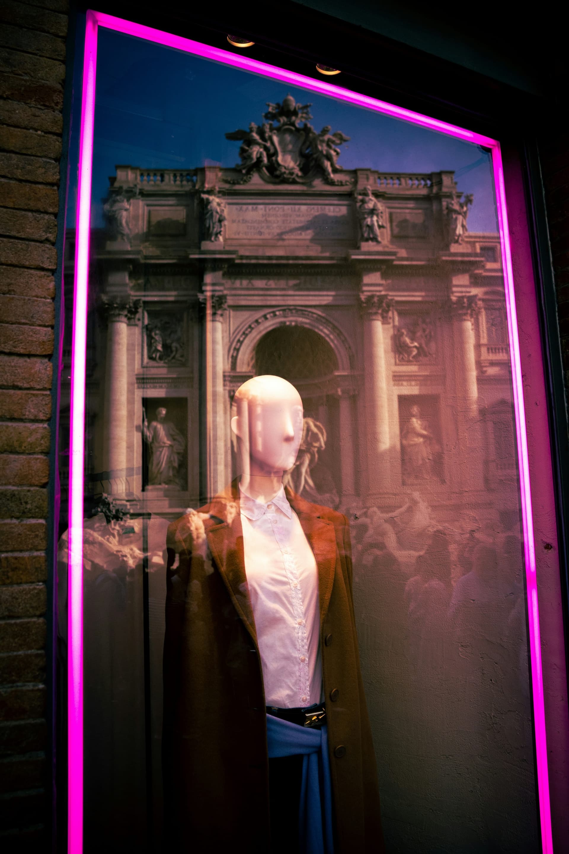 A mannequin in a white shirt and tan coat framed by pink neon, with the Trevi Fountain reflected in the glass