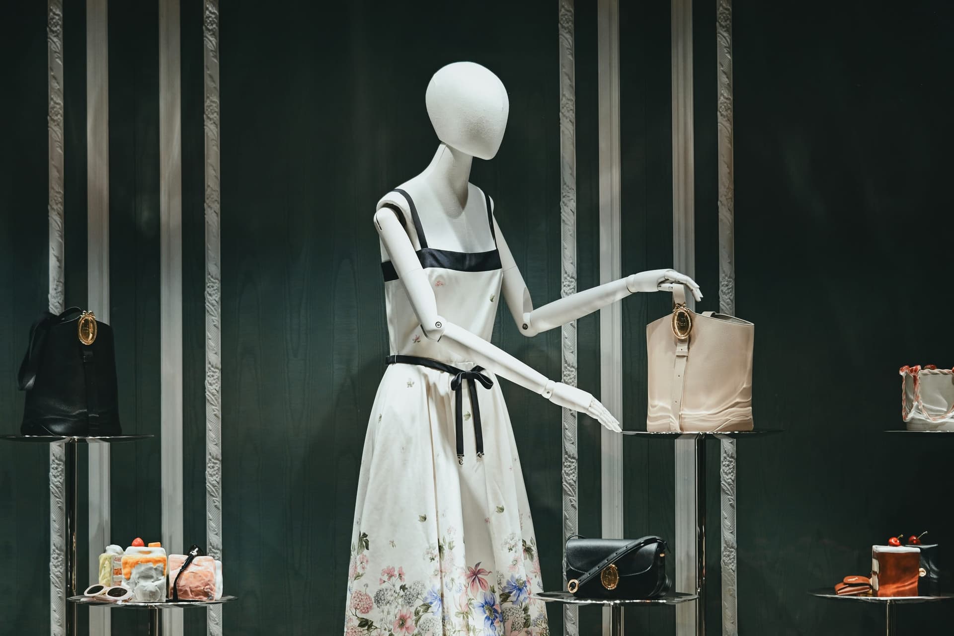 A white mannequin in a floral dress reaching for a cream bucket bag in a green paneled window